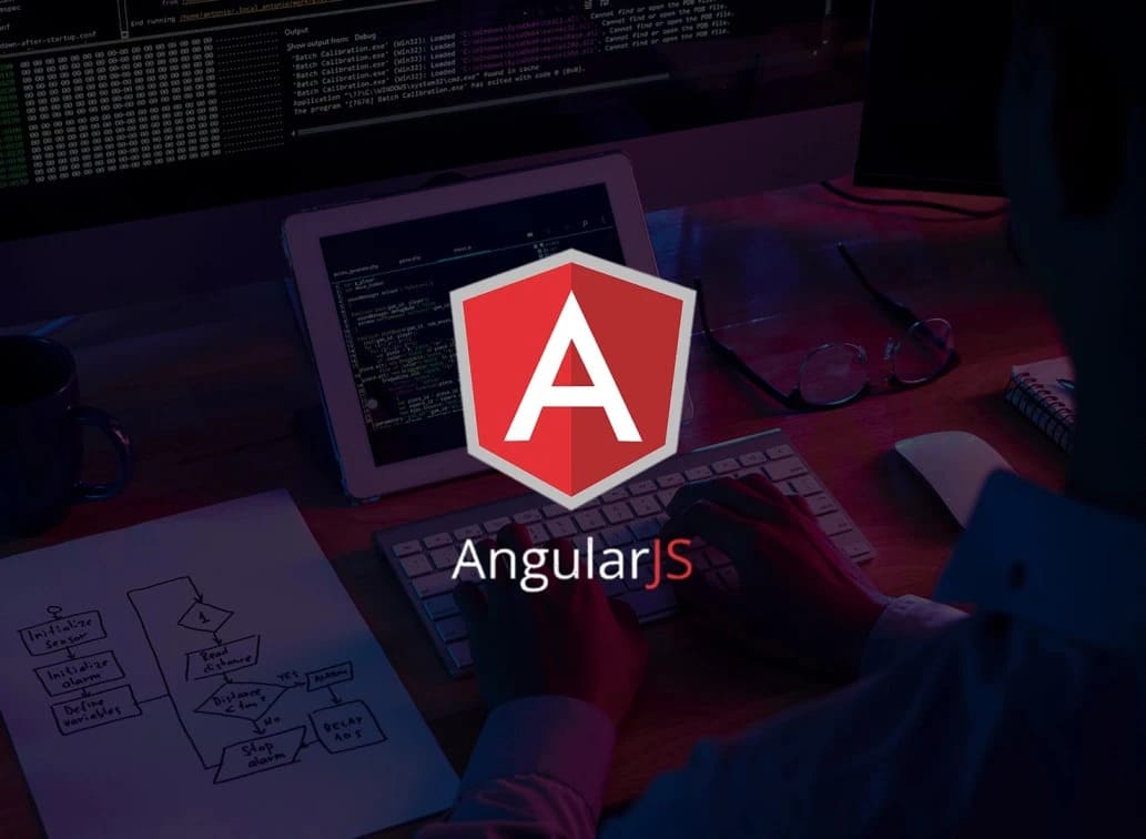 A person working at a computer with an AngularJS logo on the screen, surrounded by papers and a keyboard, in a dimly lit setting.
